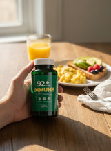 Load image into Gallery viewer, 92+ Immune Sea Moss Capsules bottle held by hand with breakfast in the background
